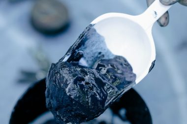 A detailed view of a hand with a spatula scooping dark mud from a jar, highlighting texture.